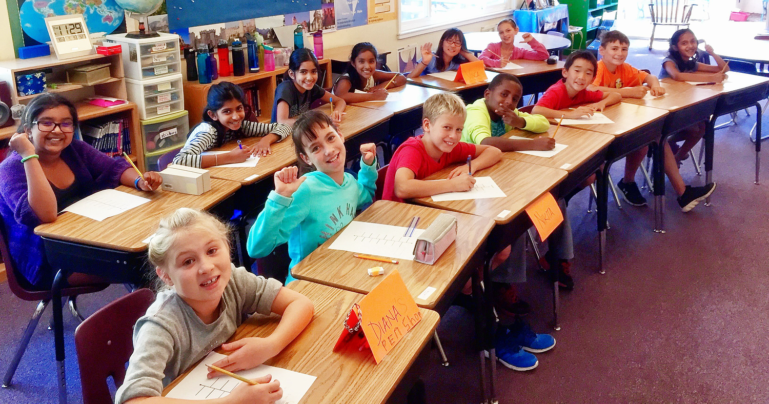 Children in 4th and 5th grades at Living Wisdom School in Palo Alto, California sit at their desks in curved rows while smiling at the photographer.