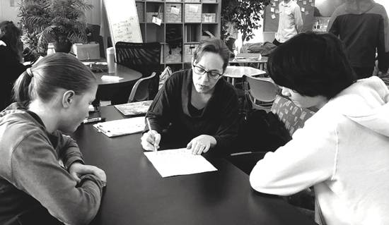 A female teacher works with students seated around a circular table at Living Wisdom High School of Palo Alto, California.