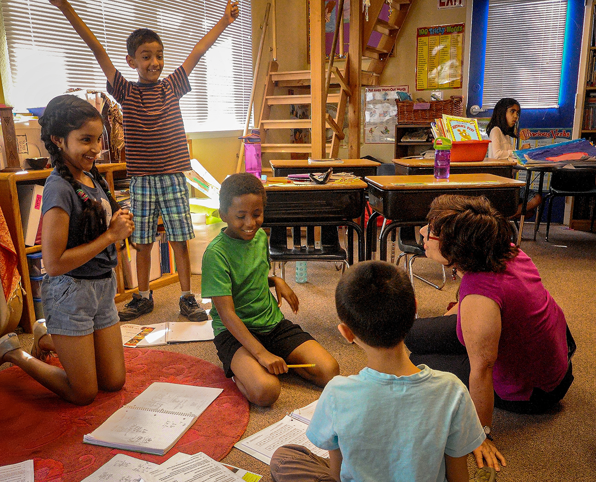 Third graders celebrate a math victory. One boy stands with arms upraised in triumph, smiling. Teacher Ruth Silver sits on floor with the other children celebrating.