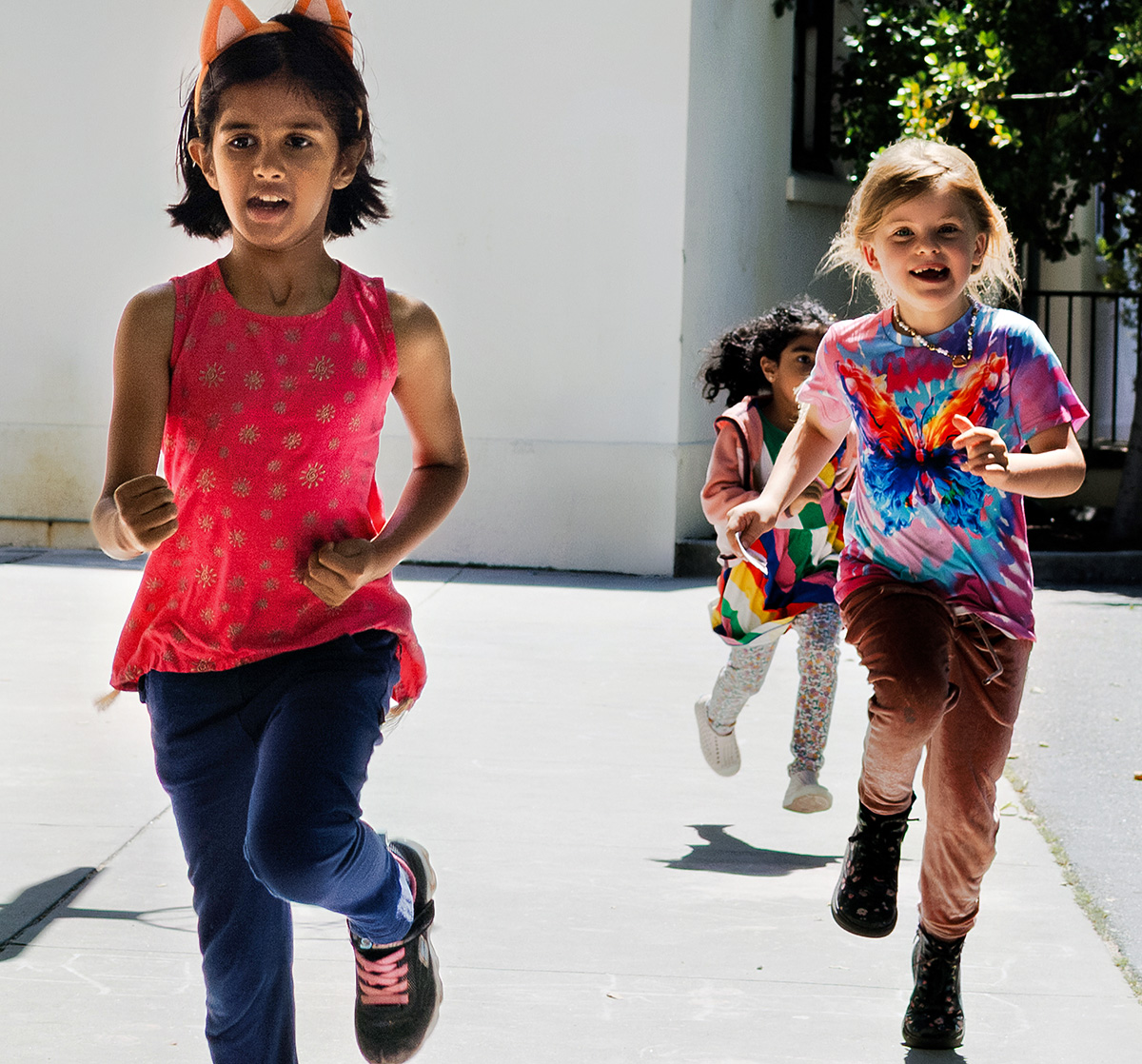Three young girls run joyfully across concrete playground in sun.